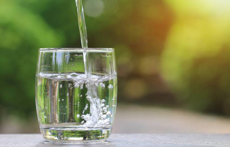 Een glas helder water waarin meer water gegoten wordt. Het water werd onthard met een CO2 waterontharder. Het glas staat op een grijze ondergrond, voor een vervaagde groene achtergrond.